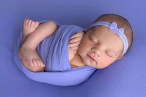 Newborn baby swaddled in purple wrap sleeping peacefully with floral headband on matching violet background.