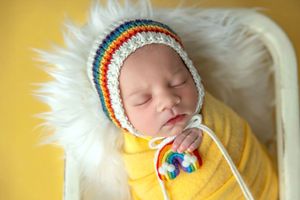 Sleeping newborn baby wrapped in yellow swaddle wearing colorful rainbow striped crochet bonnet hat on white fur.