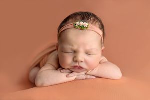 Sleeping newborn baby girl wearing a floral headband, posed with chin resting on hands against a peach background.