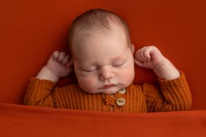 Sleeping newborn baby in orange knit cardigan on red background, peaceful infant portrait photography.