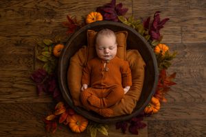Newborn baby in orange knit outfit sleeping in wooden bowl surrounded by fall leaves and mini pumpkins.