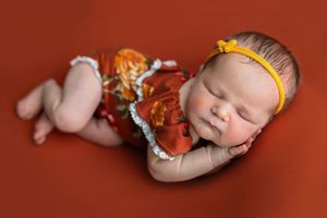 Sleeping newborn baby girl in red floral outfit and yellow headband on rust background.