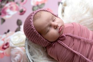 Sleeping newborn baby wrapped in dusty rose pink swaddle with knit bonnet in a white basket.
