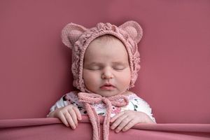 Sleeping newborn baby wearing a pink crocheted bear ear bonnet, posed on a pink background.