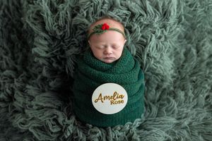 Newborn baby girl named Amelia Rose swaddled in green knit wrap with floral headband on fluffy green backdrop.