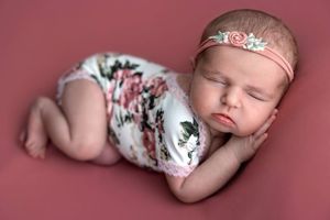 Sleeping newborn baby girl in floral romper and pink headband posed on mauve background.