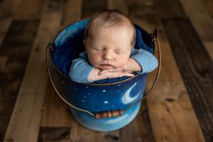 Sleeping newborn baby posed in a blue moon and stars painted metal bucket on a rustic wood floor.