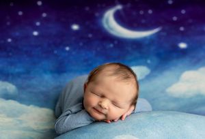 Smiling newborn baby sleeping peacefully on blue pillow with dreamy night sky moon and stars backdrop.