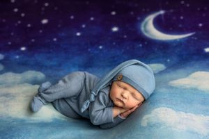 A sleeping newborn baby dressed in a blue outfit and hat, nestled among clouds under a crescent moon.
