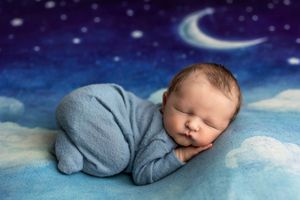 Peaceful newborn baby sleeping curled up in blue outfit on soft blue blanket with starry night sky backdrop.
