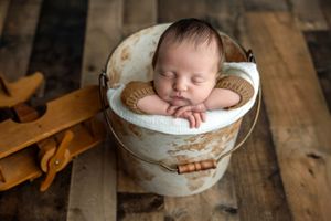 Sleeping newborn baby posed in a rustic white bucket with a wooden toy airplane on wood plank floor.