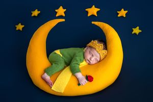Newborn baby sleeping on yellow crescent moon prop wearing green outfit and knit hat, surrounded by stars on dark blue background.