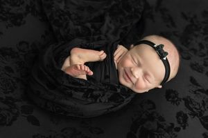 Smiling newborn baby wrapped in black floral fabric with a black bow headband, sleeping peacefully.