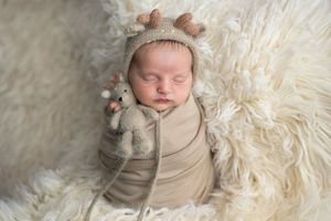 Sleeping newborn baby wrapped in beige swaddle, wearing reindeer hat, holding tiny stuffed animal on fluffy white blanket.