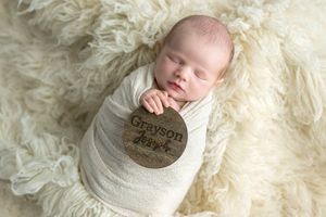 Newborn baby Grayson swaddled in white wrap sleeping on fluffy cream fur, holding a wooden name disc.