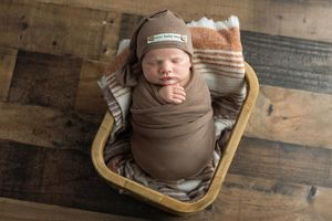 Newborn baby swaddled in brown wrap wearing a knit beanie, sleeping peacefully in a wooden basket.