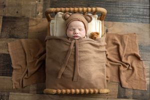 Newborn baby sleeping in tiny wooden bed wrapped in brown swaddle wearing bear ear bonnet hat.