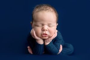 Sleeping newborn baby in blue knit outfit posed with hands under chin on navy blue background.