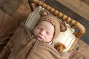 Sleeping newborn baby wearing a brown bear bonnet wrapped in a soft brown blanket in a wooden crib.