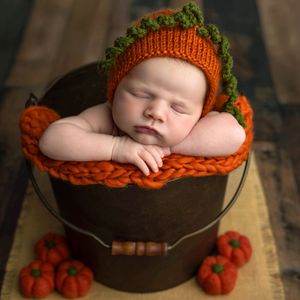 Sleeping newborn baby in orange pumpkin knit hat inside rustic metal bucket with mini pumpkins, fall-themed photo.