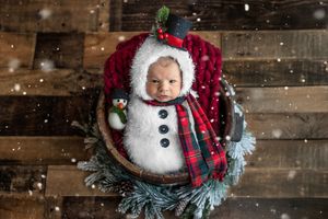 Newborn baby dressed in a snowman costume with top hat and plaid scarf, nestled in a wooden bowl with Christmas decor.