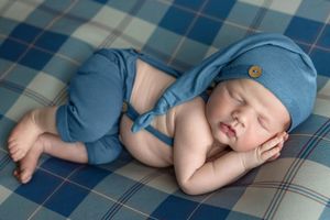Sleeping newborn baby in blue hat and suspenders outfit lying on plaid blanket.