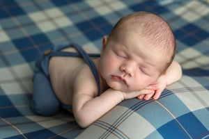 Newborn baby sleeping peacefully on a blue plaid blanket, resting chin on hands in a cute posed portrait.