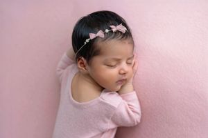 Sleeping newborn baby girl in pink outfit and bow headband on pink background.