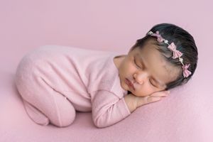 Newborn baby girl sleeping peacefully in pink outfit and floral headband on soft pink blanket.