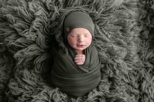 Newborn baby swaddled in olive green wrap and matching hat, sleeping peacefully on a fluffy gray textured blanket.