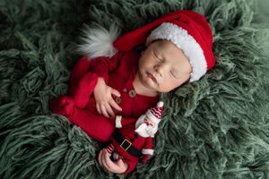 Newborn baby sleeping in red Santa Claus outfit and hat on green fur blanket with tiny Santa doll.