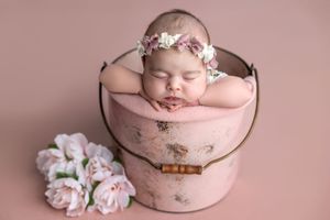 Sleeping newborn baby girl in pink floral headband resting in a vintage pink bucket with peonies.