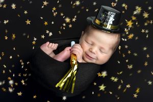 Smiling newborn baby wrapped in black with tiny top hat surrounded by gold stars for New Year celebration photo.