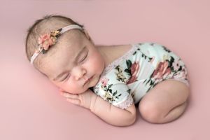 Sleeping newborn baby girl in floral romper and pink flower headband on pink backdrop.