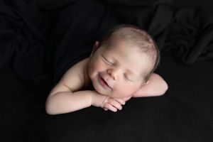 Smiling newborn baby sleeping peacefully on a black fabric backdrop with hands tucked under chin.
