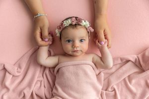 Newborn baby girl in pink wrap with floral headband lying on pink blanket held by mother's hands.