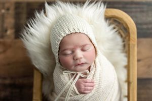 Sleeping newborn baby wrapped in cream knit swaddle wearing matching bonnet in a fluffy white fur basket.