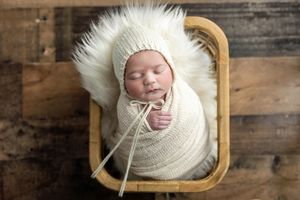 Newborn baby swaddled in cream knit wrap wearing fluffy bonnet, sleeping peacefully in a wooden basket.