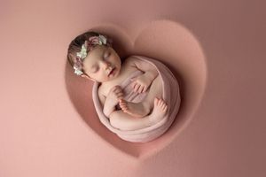 Sleeping newborn baby girl wrapped in pink, wearing floral headband, posed in heart-shaped prop for newborn photography.