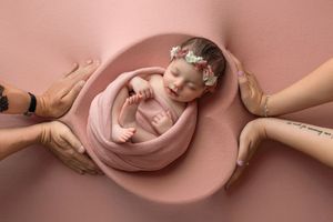 Newborn baby girl wrapped in pink swaddle resting in bowl, surrounded by parents hands, floral headband.