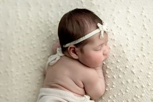 Sleeping newborn baby girl with dark hair wearing a delicate white floral headband, wrapped in cream fabric.