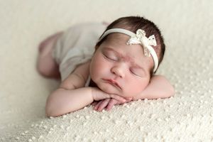 Peaceful newborn baby girl sleeping on a cream blanket, wearing a white bow headband, in a classic posed newborn photo.