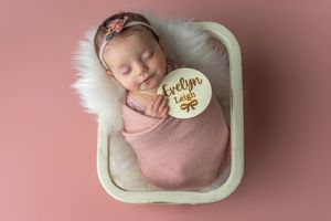 Newborn baby girl named Evelyn Leigh swaddled in pink wrap, sleeping peacefully in a cream basket on pink background.