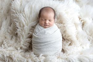 A peaceful newborn baby swaddled in a white knit wrap, resting on a fluffy cream fur blanket.