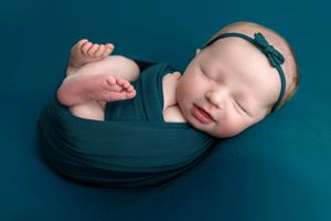 Sleeping newborn baby wrapped in teal swaddle with bow headband on teal background.