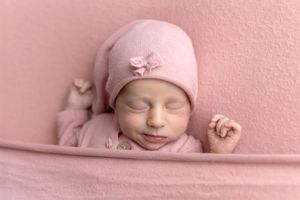 Peaceful newborn baby girl sleeping in pink outfit and hat on soft pink blanket background.