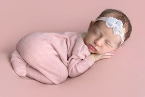 Sleeping newborn baby girl wrapped in pink swaddle with white flower headband on pink background.