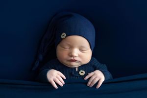 Sleeping newborn baby in navy blue knit romper and slouchy hat posed on dark blue fabric backdrop.