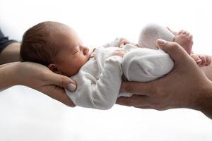 Newborn baby sleeping peacefully in parents hands wearing white onesie against light background.