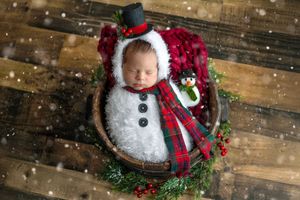 Newborn baby dressed as a snowman in a basket with holiday greenery and plaid scarf, with falling snow effect.